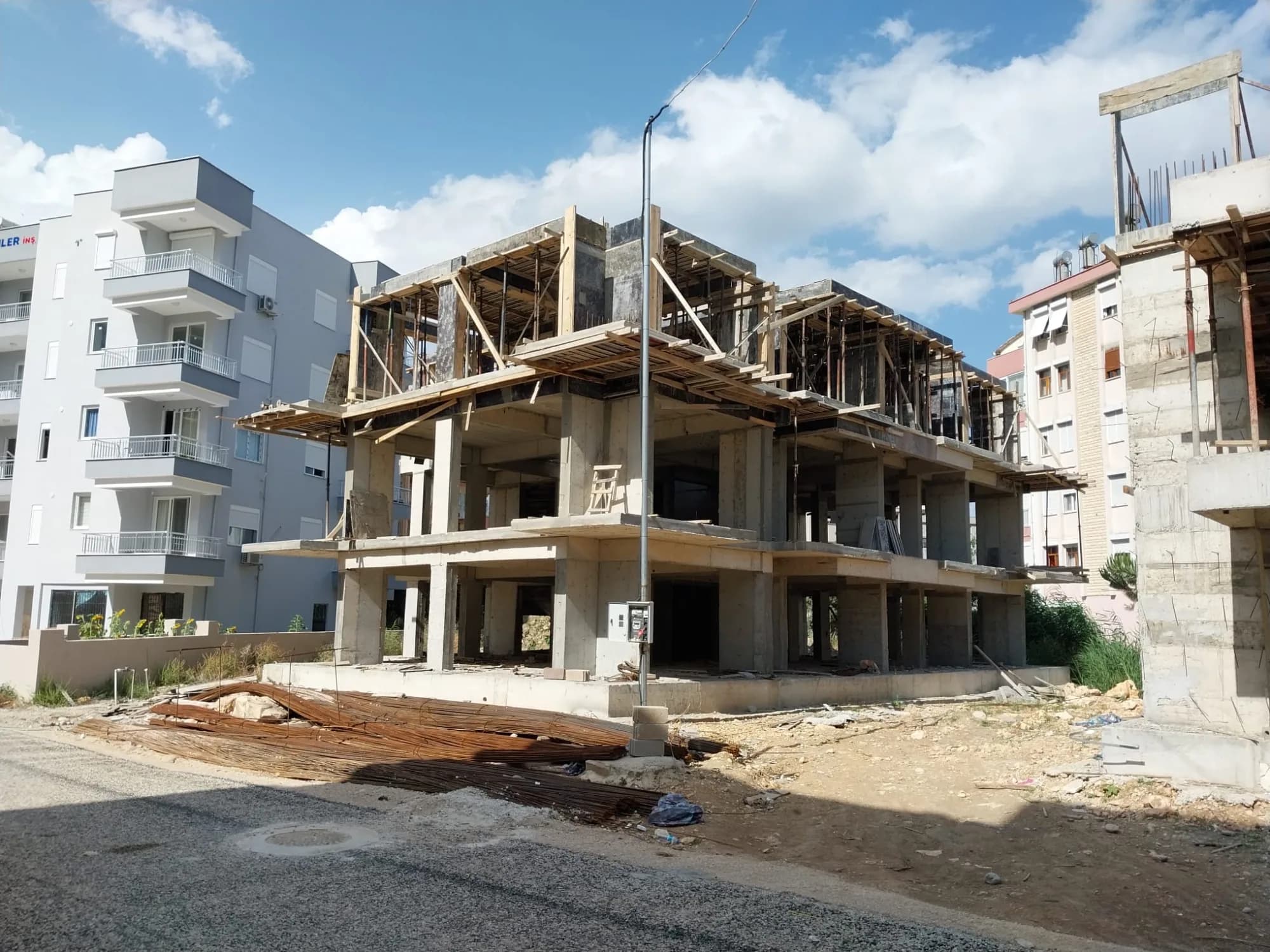 Dönüşüm öncesi: Finike'de kaba inşaat hâlindeki bina; kalıpları sökülmemiş betonarme iskelet, sahada istiflenmiş inşaat demirleri.